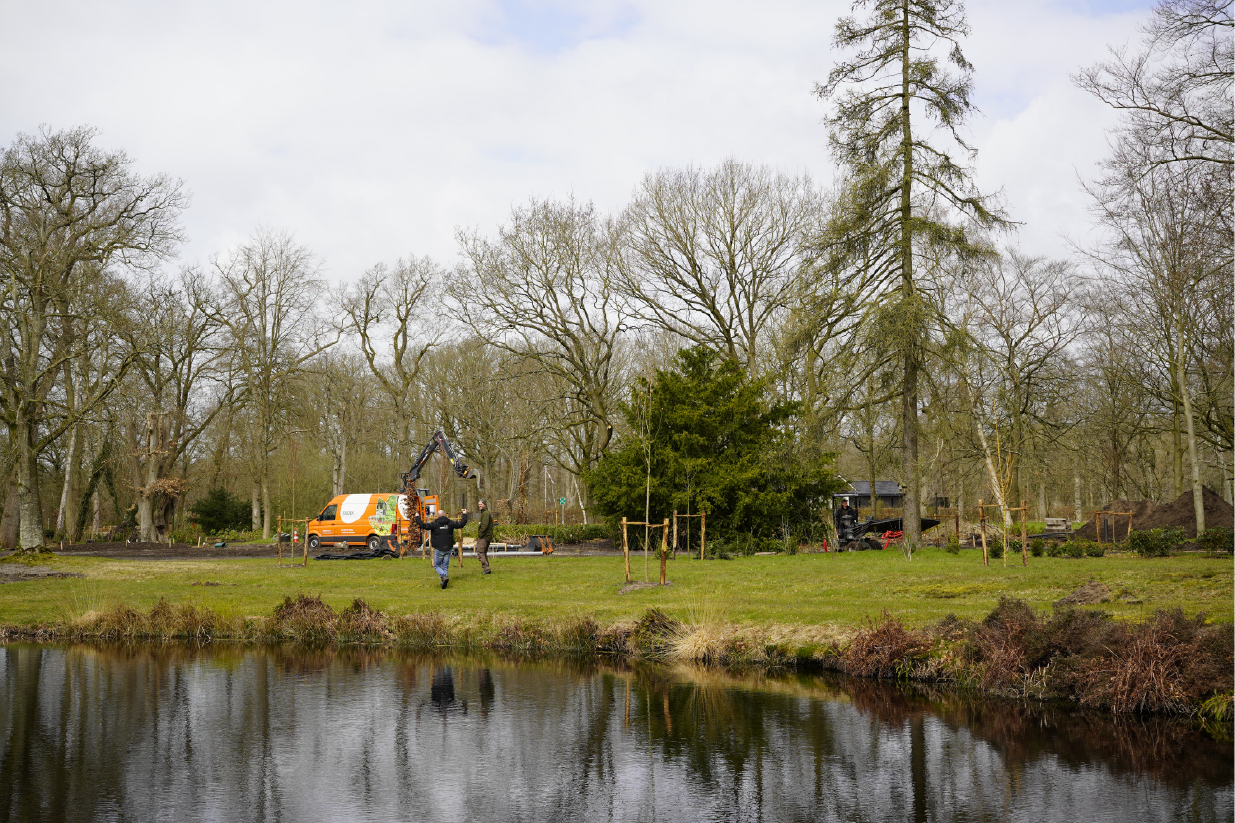 Op de voorgrond een vijver met in de verte een oranje Snoek Puur Groen bus en twee mensen met elkaar in gesprek. Daarachter bomen.
